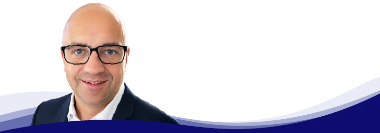 Tom Ruegenberg, Projektmanager bei Gebr. Kemper, lächelt und blickt in die Kamera. Er trägt eine Brille mit schwarzem Gestell, ein hellblaues Hemd mit offenem Kragen und ein graues Sakko darüber. Er hat eine Glatze und ist glatt rasiert.