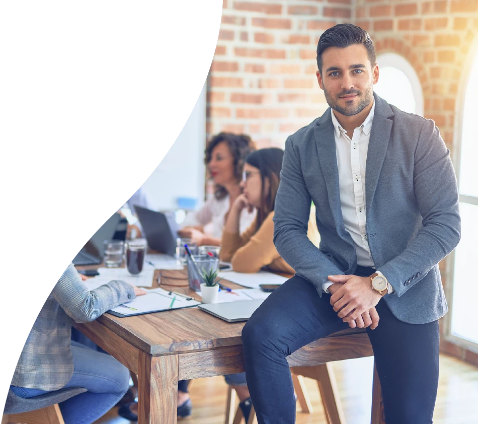 Ein junger Mann in Business-Outfit sitzt am Rand eines Konferenztisches und blickt direkt in die Kamera. Im Hintergrund arbeiten mehrere Personen konzentriert zusammen. Die Szene spielt in einem modernen Büro mit Ziegelwand.