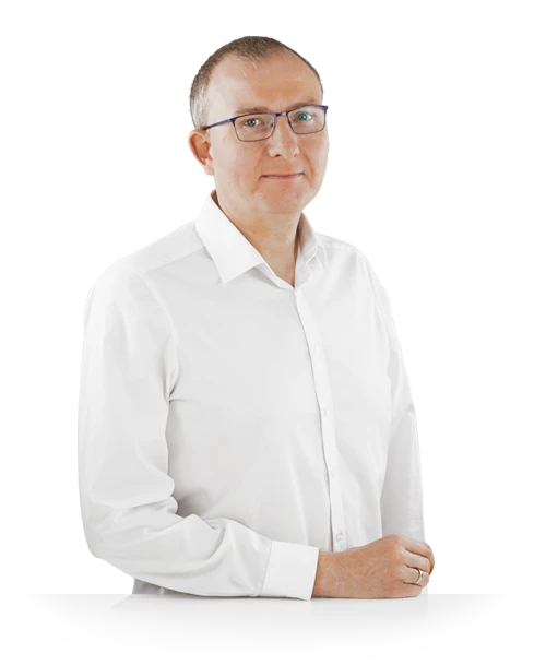 Johannes Gonser steht mit weißem Hemd hinter einem Tisch. Er steht seitlich, die rechte Hand entspannt auf der Tischplatte, und blickt mit einem angedeuteten Lächeln in die Kamera.
