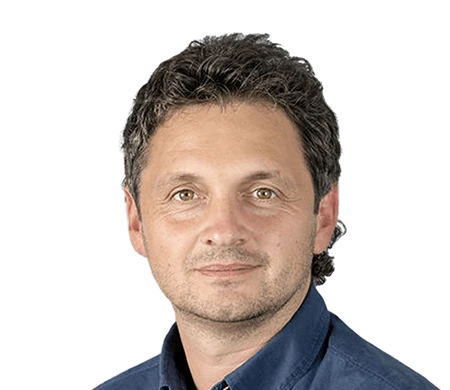 Reinhard Sutterlüty der Firma meusburger lächelt in die Kamera. Er hat lockiges, dunkles Haar und trägt ein dunkelblaues Hemd. Der Hintergrund zeigt eine moderne Wellenform in Weiß, Hellblau und Dunkelblau, die sich geschwungen von links unten nach rechts oben über das Bild zieht.