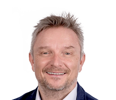 Holger Geck, Head of Marketing and Product Data Management at Gust. Alberts, smiles into the camera. He has short, gray-blond hair and a short beard. He is wearing a white shirt and a dark jacket. A curved wave shape in white, light blue and dark blue can be seen in the background.