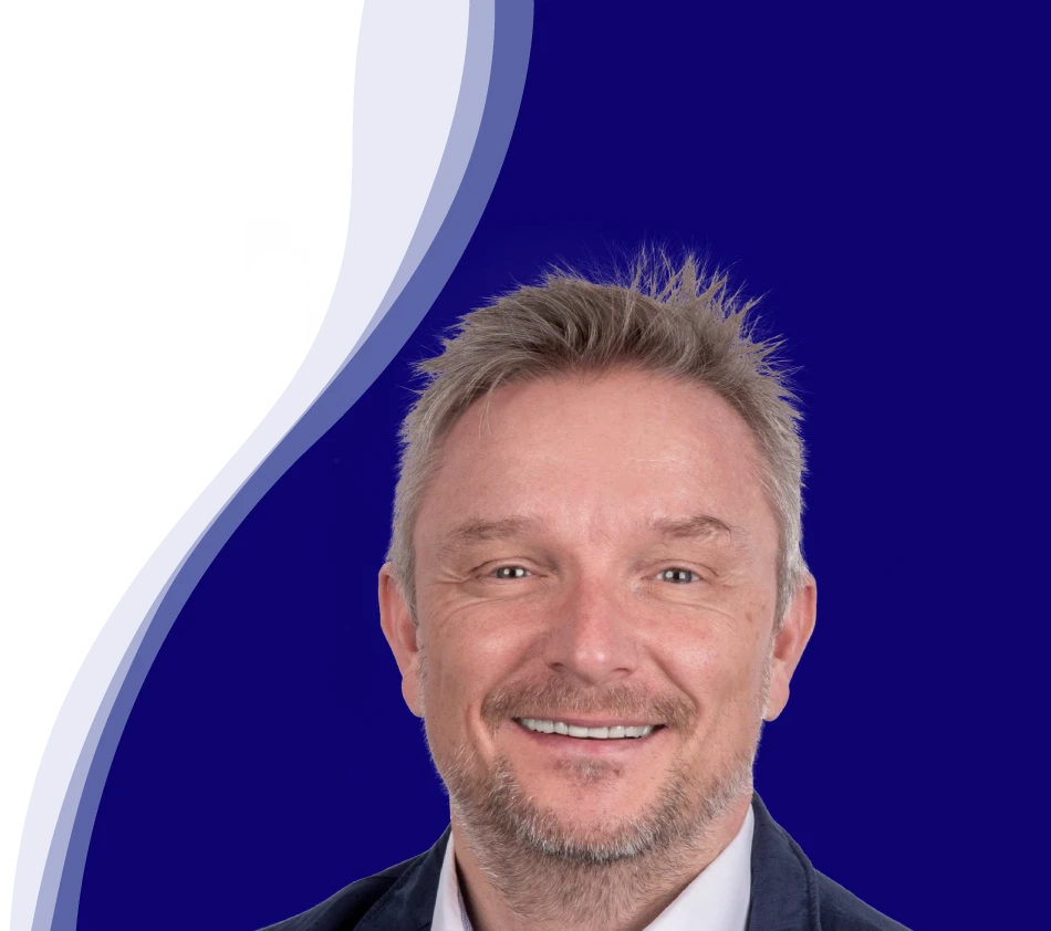 Holger Geck, Head of Marketing and Product Data Management at Gust. Alberts, smiles into the camera. He has short, gray-blond hair and a short beard. He is wearing a white shirt and a dark jacket. A curved wave shape in white, light blue and dark blue can be seen in the background.