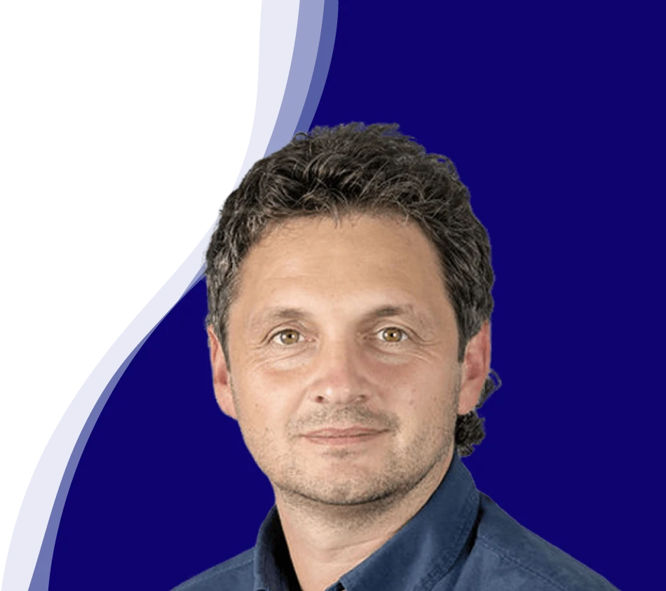 Reinhard Sutterlüty from meusburger smiles at the camera. He has curly, dark hair and is wearing a dark blue shirt. The background shows a modern wave shape in white, light blue and dark blue, which curves from the bottom left to the top right of the picture.