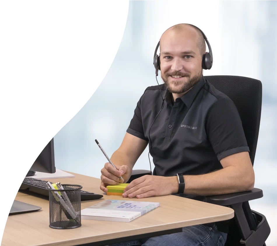 Un collaborateur crossbase du service de conseil regarde la caméra en souriant. Il est assis à son bureau, un casque sur la tête, un stylo et un bloc-notes à la main.