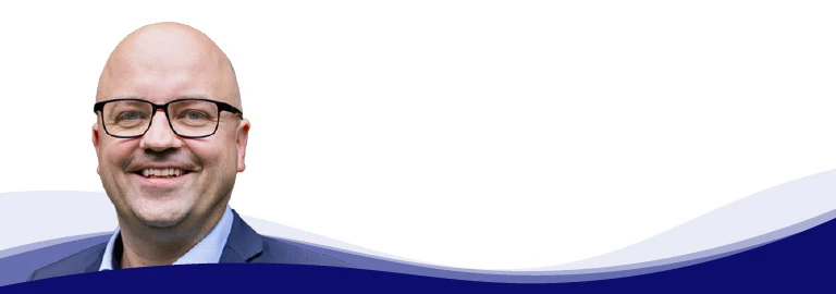 Tom Ruegenberg, chef de projet chez Gebr. Kemper, sourit et regarde la caméra. Il porte des lunettes à monture noire, une chemise bleu clair à col ouvert et une veste grise par-dessus. Il est chauve et rasé de près.